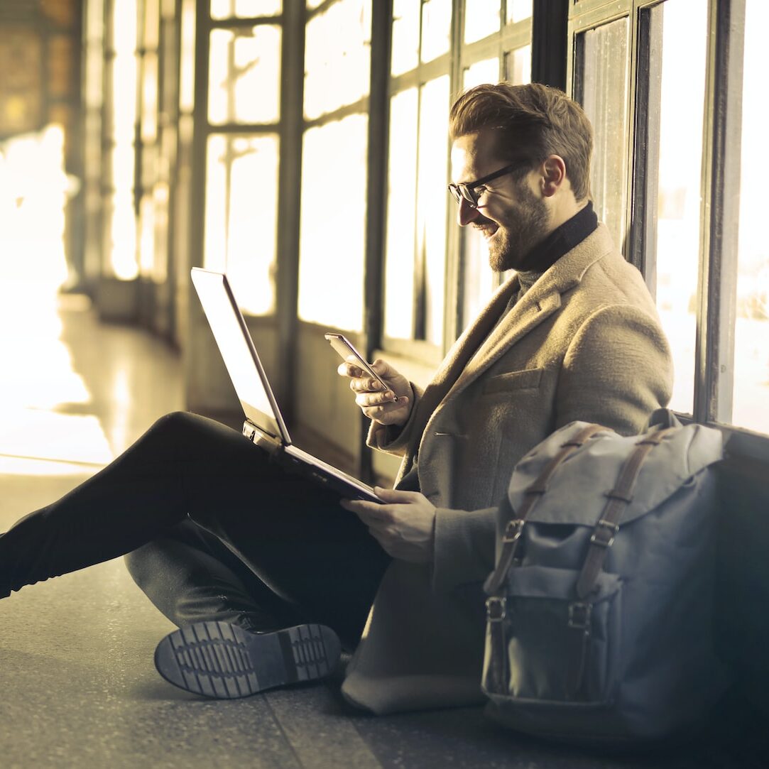 man sitting near window holding phone and laptop